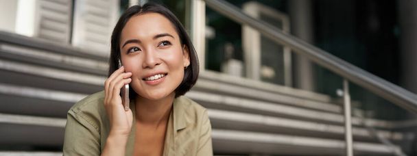 Portrait of asian girl smiles while talks on mobile phone. Young woman calling a friend, sitting on stairs. Technology concept - Photo, Image