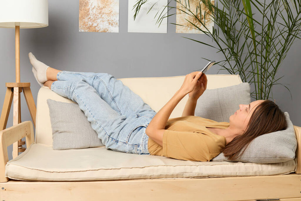 Tech addiction. Smartphone browsing. Digital leisure. Wireless connection. Internet relaxation. Brown haired woman wearing beige T-shirt lying on sofa holding smartphone in living room - Фото, изображение