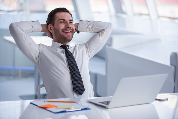Hombre de negocios guapo en traje y auriculares está sonriendo mientras trabaja con el ordenador portátil en la oficina
 - Foto, Imagen