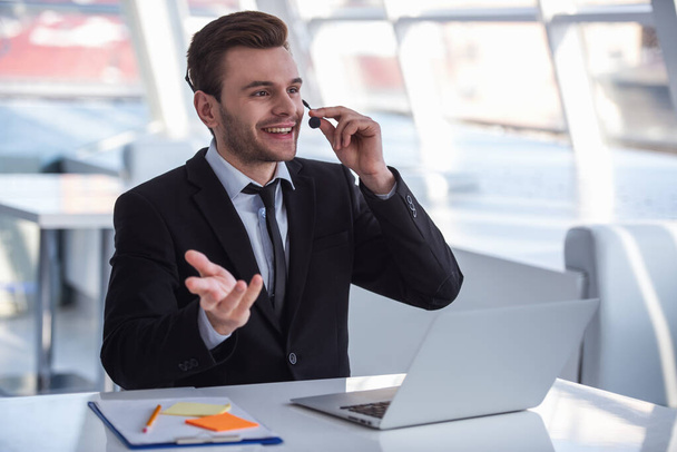 Привлекательный бизнесмен в костюме и гарнитуре разговаривает и улыбается во время работы с ноутбуком в офисе
 - Фото, изображение