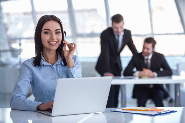 attraktive Geschäftsfrau mit Headset lächelt, während sie im Büro mit Laptop arbeitet, ihre Kollegen im Hintergrund - Foto, Bild
