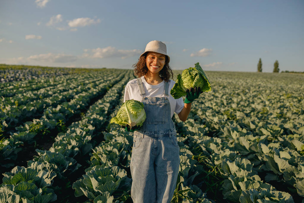 Χαμογελαστή αγρότισσα με στολή που εργάζεται στον τομέα του λάχανου κατά τη συγκομιδή. Γεωργική δραστηριότητα  - Φωτογραφία, εικόνα