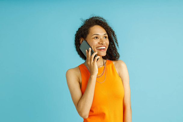 Csinos nő narancssárga ruhában beszél mobiltelefonnal, miközben áll a kék stúdió háttér - Fotó, kép