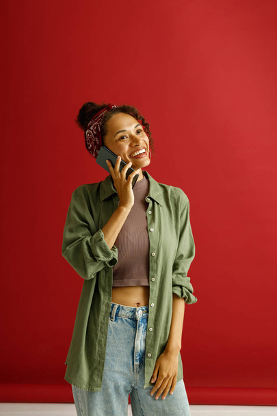 Pretty woman in casual clothes is talking by mobile phone while standing on red studio background - Fotoğraf, Görsel