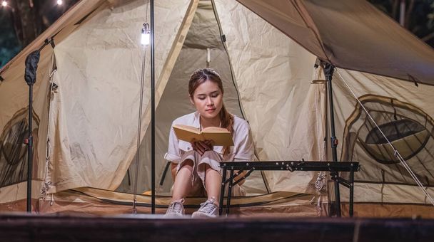 Νεαρή τουρίστρια στα 30 της με ρούχα σαφάρι κάθεται σε μια σκηνή κάμπινγκ και διαβάζοντας ένα βιβλίο, υπαίθρια ταξιδιωτική δραστηριότητα Χαλαρώστε μέσα στη φύση. - Φωτογραφία, εικόνα