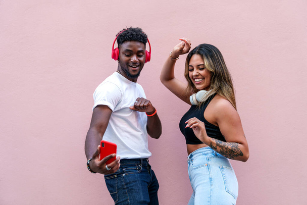 Lachende, stijlvolle stedelijke modellen van diverse etniciteiten dansen op muziek via oortelefoons tegen een roze achtergrond. - Foto, afbeelding