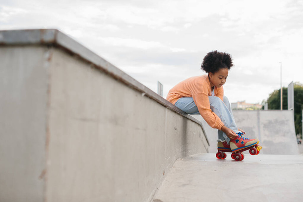 Cute African American teen girl putting on roller skates while sitting on a ramp at a skatepark on the beach. - Foto, Imagen