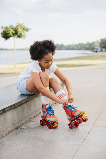 Cute African American teen girl putting on roller skates while sitting on a ramp at a skatepark on the beach. - Photo, Image