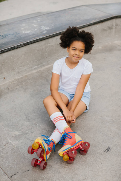 Carino afro americano ragazza pattinatore seduto nel parco della città e sorridente. - Foto, immagini