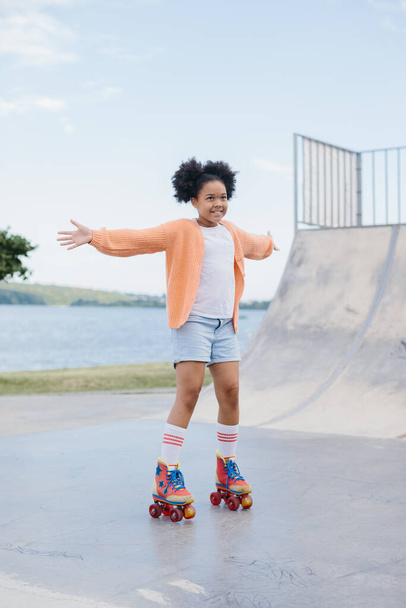 Petite fille afro-américaine mignonne chevauchant sur patins à roulettes dans un parc d'été. Enfant jouissant de sport en plein air dans le parc, drôle petite fille jouant sur le patin à roulettes sur la route. sport apprentissage concept outdoor - Photo, image