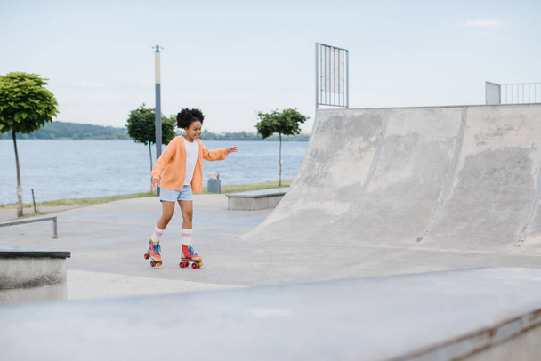 Мила афроамериканська маленька дівчинка катається на роликових ковзанах в літньому парку. Малюк насолоджується спортом на відкритому повітрі в парку, смішна дівчинка грає на роликових ковзанах на дорозі. спортивне навчання на відкритому повітрі концепція - Фото, зображення