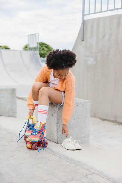 A teenage girl takes off her roller skates after skating. African American girl having fun at the skatepark on the weekend. - Photo, Image