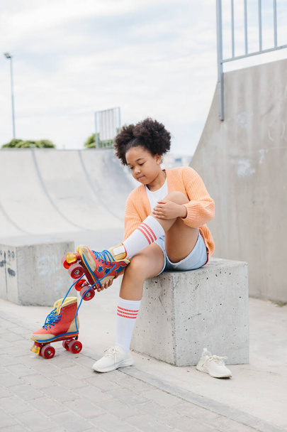 A teenage girl takes off her roller skates after skating. African American girl having fun at the skatepark on the weekend. - Photo, Image