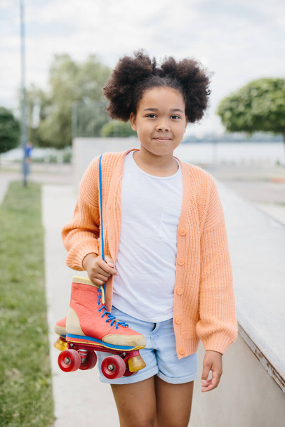 A young girl came to skate in the skatepark. African American girl holding vintage rollers tied with shoelaces. - Photo, Image