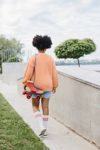 Una joven vino a patinar en el parque de skate. Chica afroamericana sosteniendo rodillos vintage atados con cordones. - Foto, Imagen