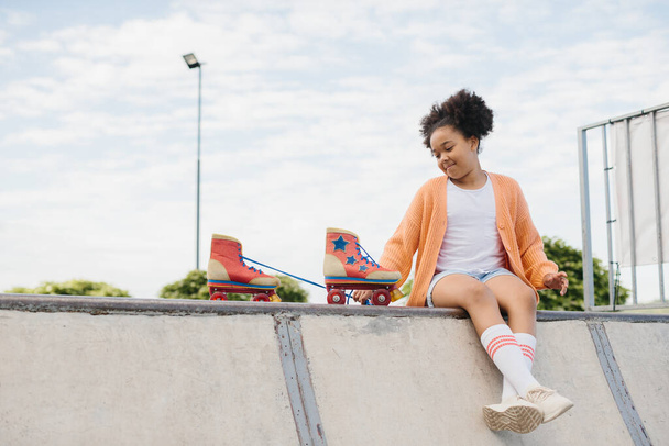 Adolescente noire africaine active assise en train de se détendre avec ses patins à roulettes au bord de la plage, souriant pendant une journée ensoleillée d'été en vacances, en plein air. Fille joyeuse attrayant contre un ciel bleu. - Photo, image
