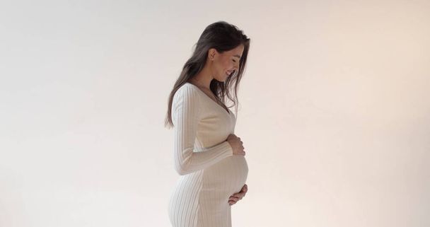 Schöne schwangere Mutter posiert im Studio auf weißem Hintergrund. Eine junge brünette schwangere Frau in einem weißen Kleid streichelt ihren Bauch und genießt ihre Schwangerschaftsphase. - Foto, Bild