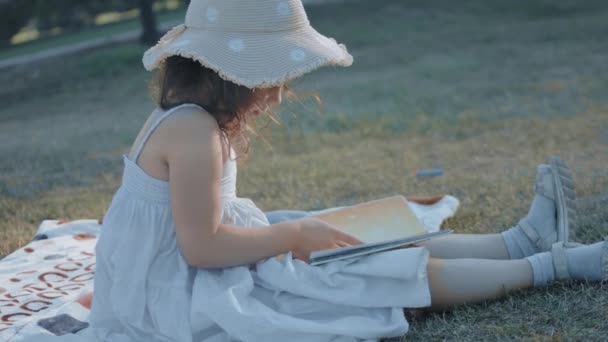 Jolie petite fille bouclée portant un chapeau de soleil assis sur une couverture et lit un livre lors d'un pique-nique dans la nature. Concept d'été, vacances, loisirs et enfance heureuse - Séquence, vidéo