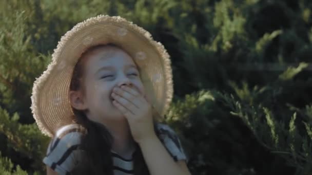 Jolie petite fille en robe rayée et chapeau de soleil relaxant sur l'herbe dans le parc. Rire d'une blague. Été, vacances, concept d'enfance heureuse. Gros plan. - Séquence, vidéo