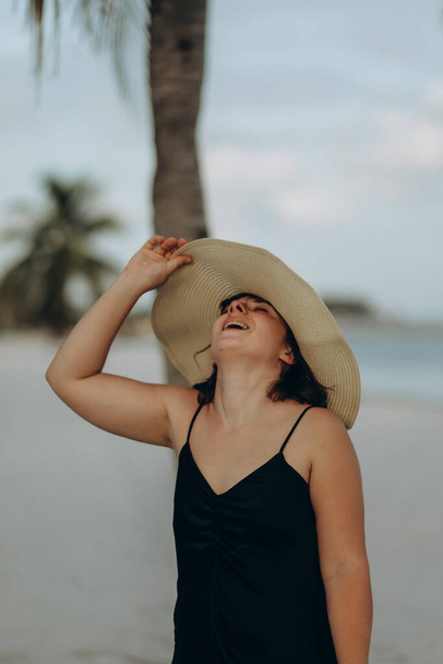 Дівчина в чорній сукні в солом'яному капелюсі на березі океану з пальмами - Фото, зображення