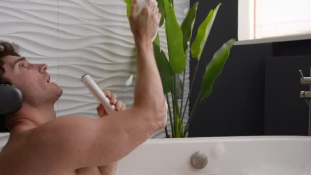 泡でリラックスする白人男性 遊び心のある幸せな男性 ヘッドフォン付き シャワージェルボトルシャンプーで音楽を聞く 朝の浴室でオーディオリラックス - 映像、動画