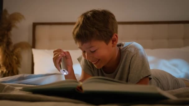 Gülümseyen küçük beyaz çocuk okul çocuğu elinde fenerle yatak odasında kitap okuyor. Gece boyunca masal okuyup öğrenmenin keyfini çıkarıyor. - Video, Çekim
