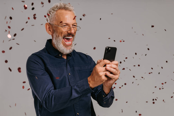 Happy mature man looking at smart phone with confetti flying around on gray background - Фото, зображення