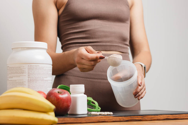 Athletic woman in sportswear with measuring spoon in her hand puts portion of whey protein powder into a shaker on wooden table with amino acid white capsules, bananas and apple, making protein drink. - Photo, Image