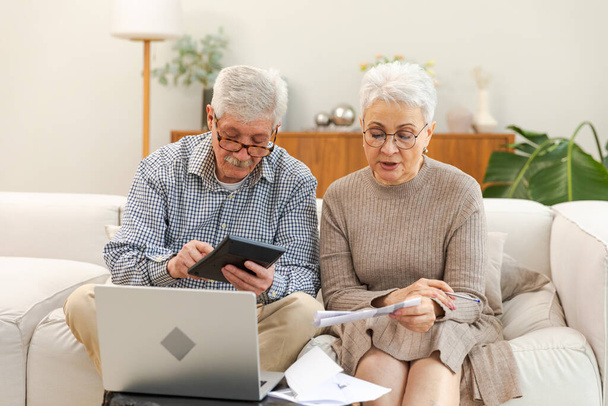 Middle aged senior couple sit with laptop and paper document. Older mature man woman reading paper bill pay online at home managing bank finances calculating taxes planning loan debt pension payment - Photo, Image
