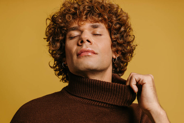 Fashionable krullende jongeman stretching trui nek en houd de ogen gesloten tegen een gele achtergrond - Foto, afbeelding