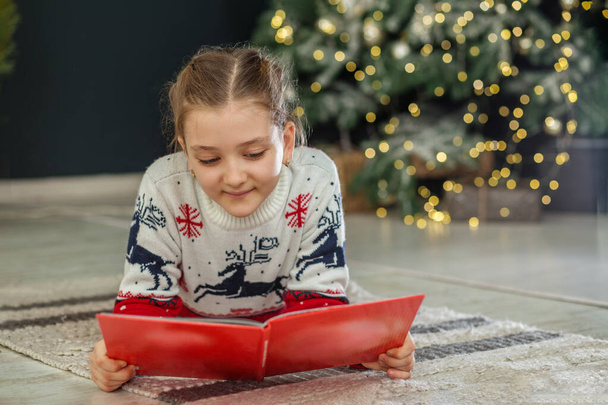 Ένα χαμογελαστό παιδί διαβάζει βιβλίο στο δωμάτιο. Έννοια Πρωτοχρονιά, Καλά Χριστούγεννα, διακοπές, διακοπές, χειμώνα, παιδική ηλικία. Άνετο σπίτι.. - Φωτογραφία, εικόνα