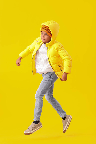 Jumping African-American boy in warm winter clothes on yellow background - Photo, Image