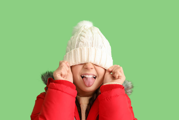 Смешная маленькая девочка в шляпе и тёплая куртка на зелёном фоне, крупный план - Фото, изображение