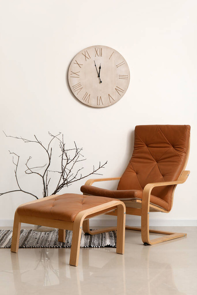 Άνετο καφέ πολυθρόνα με πόδι σκαμνί, κλαδιά δέντρων στο βάζο και ρολόι κοντά στο λευκό τοίχο - Φωτογραφία, εικόνα