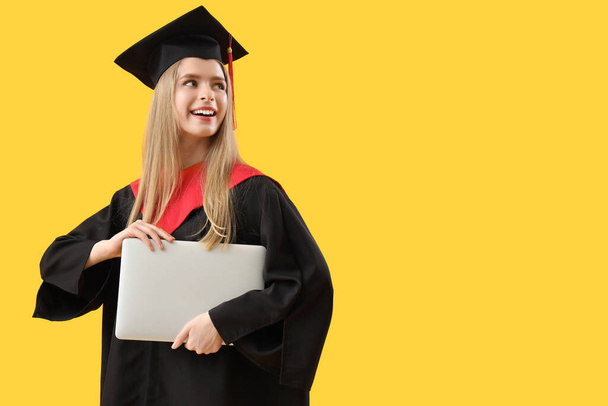 Vrouwelijke afgestudeerde student met laptop op gele achtergrond - Foto, afbeelding