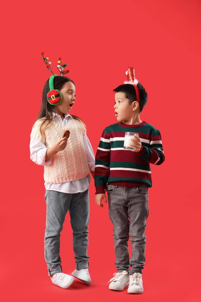 Surprised little children with Christmas decor, glass of milk and cookie on red background - Photo, Image