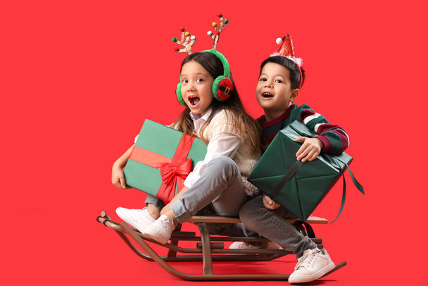Emotional little children with Christmas gifts and sleigh on red background - Photo, Image