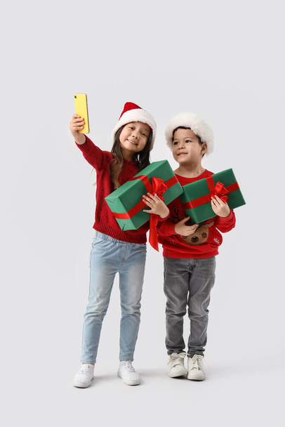Cute little children with Christmas gifts taking selfie on light background - Photo, Image