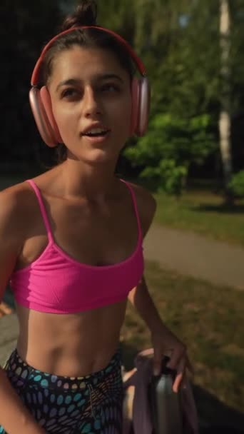 Uma jovem encantadora em trajes atléticos e fones de ouvido está saciando sua sede enquanto treina ao ar livre. Imagens 4k de alta qualidade - Filmagem, Vídeo