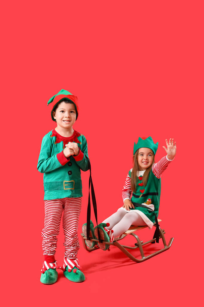 Cute little children dressed as elves with sledge on red background - Photo, Image