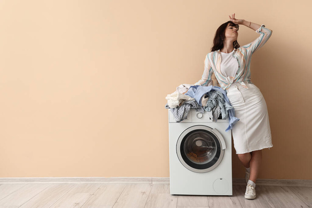 Κουρασμένη νεαρή γυναίκα με βρώμικα ρούχα και πλυντήριο κοντά σε μπεζ τοίχο - Φωτογραφία, εικόνα