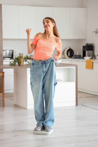 Happy beautiful young woman in loose jeans with scales at kitchen. Weight loss concept - Фото, изображение