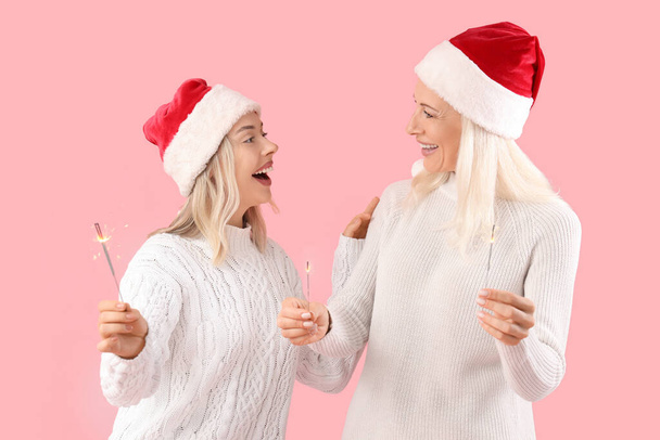 Happy daughter and her mother in Santa hats with sparklers on pink background - Photo, Image