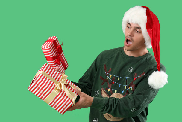 Yeşil arka planda hediye kutularıyla Noel Baba şapkalı genç adam şok oldu. - Fotoğraf, Görsel