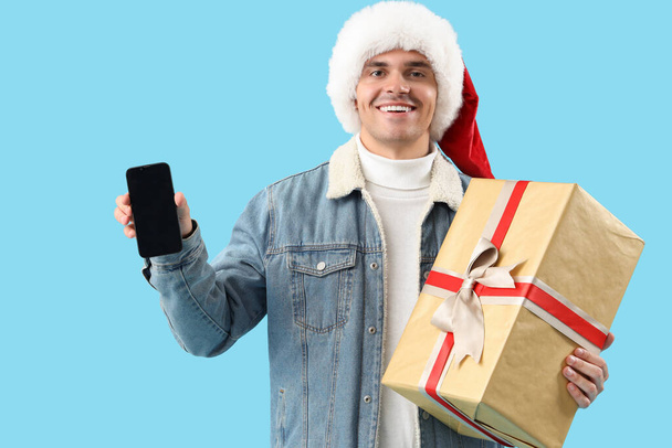 Happy young man in Santa hat with gift box and mobile phone on blue background - Photo, Image
