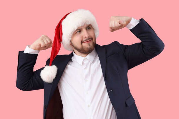 Junger Mann mit Weihnachtsmütze zeigt Muskeln auf rosa Hintergrund - Foto, Bild
