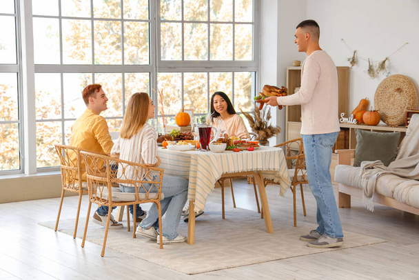 Jovem trazendo saboroso peru à mesa de jantar com seus amigos em casa no Dia de Ação de Graças - Foto, Imagem