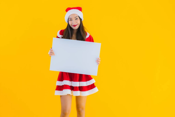 Portrét krásné mladé asijské ženy vánoční oblečení a klobouk úsměv s prázdnou bílou tabuli znamení na žlutém izolovaném pozadí - Fotografie, Obrázek