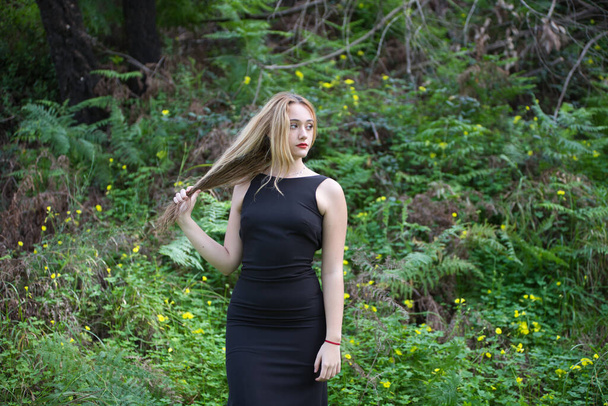 Giovane, bella donna bionda vestita di nero cammina attraverso la foresta in diverse posture ed espressioni. Sullo sfondo felci e fiori gialli. Espressioni concettuali in natura. - Foto, immagini
