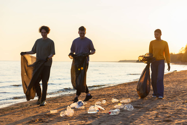 Dzień Ziemi. Zespół aktywistów wolontariuszy zbiera śmieci w strefie przybrzeżnej plaży. Kobieta ze śmieciami w worku na śmieci na brzegu oceanu. Ochrona środowiska czyszczenie stref przybrzeżnych - Zdjęcie, obraz